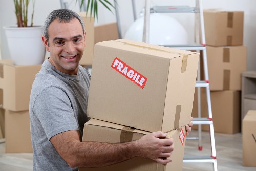 A cheerful man with short, dark hair and a light complexion is standing indoors in a residential setting, holding two cardboard boxes stacked one on top of the other. The top box has a red and white 'FRAGILE' label. He is wearing a casual grey t-shirt and smiling directly at the camera. Behind him, there is a stainless steel ladder and several other cardboard boxes of various sizes, some sealed with packing tape, arranged around the room. A white potted plant with broad green leaves is visible on the left, and the background includes a neutral-toned wall, suggesting an organized space possibly used for packing or storage before moving. The scene appears well-lit with natural or bright artificial lighting, consistent with a house or flat in Tooting, London, as part of professional removals services provided by Tooting Man with Van, specializing in house relocations and move logistics.