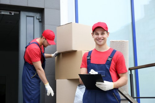 A professional house removals team in Tooting, London, preparing for a residential move outside a modern building with a dark exterior wall and large glass windows. One mover, smiling at the camera, is wearing a red cap, red t-shirt, and navy blue overalls, holding a clipboard and standing near several stacked cardboard boxes of varying sizes, some wrapped in packing tape. The second mover, also dressed in a red cap and navy blue overalls with a red t-shirt, is focused on lifting or adjusting a large cardboard box from the ground, with additional boxes visible behind him. The scene is set in natural daylight, indicating daytime, and shows the movers on a paved pathway or driveway adjacent to the building's entrance. This image, representative of local removals services, highlights the careful handling of household items and professional staff ready to assist with house relocations in the Tooting area.