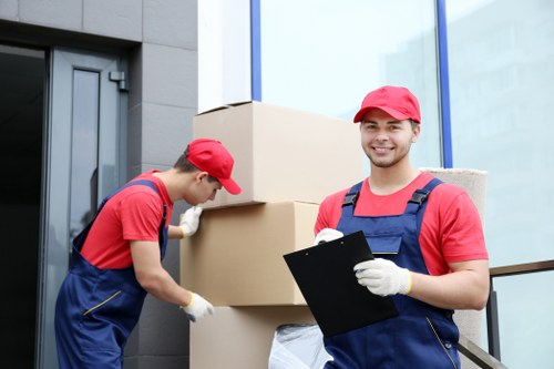 Two professional movers from Tooting Man with Van, dressed in red caps, red shirts, and dark blue overalls, are handling large cardboard moving boxes outside a modern residential building. The mover on the right is holding a black clipboard and smiling at the camera, while the other is focused on securing a box. The boxes, made of brown cardboard, vary in size and are stacked on top of each other, ready for transportation. The background features the exterior of a contemporary house with grey walls, a glass door, and large windows. The scene is illuminated by natural daylight, suggesting a clear day in the Tooting area, part of south London, reflecting typical house removals activity. The environment indicates an organized move from a residential property, with the focus on careful handling and professional service, consistent with local removals for homes in the postcode area.
