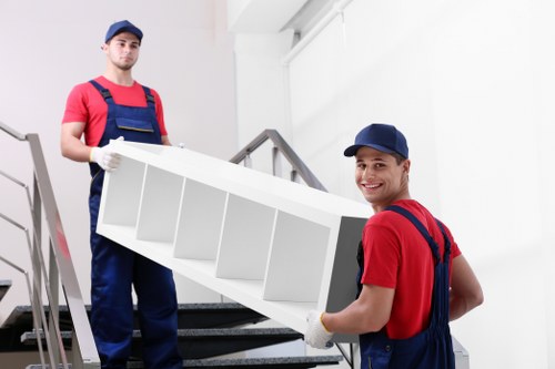 Two professional removalists wearing matching red shirts, navy blue overalls, and protective gloves are lifting a white wooden bookshelf with multiple compartments, preparing to move it up a staircase inside a modern residential property. The setting appears to be an interior with a white wall, metal staircase railing, and a carpeted staircase leading to an upper floor. The removalists are focused and smiling, demonstrating careful handling of the furniture. Natural lighting illuminates the scene, highlighting the textures of the wood and fabric of their uniforms. In the background, the staircase's dark carpet contrasts with the light-colored wall, and the staircase is narrow, suggesting the move is within a typical London or Tooting residential property. This scene reflects professional house removal services in the Tooting area, emphasizing careful and efficient handling of household furniture during a move, coordinated by the company Tooting Man with Van.