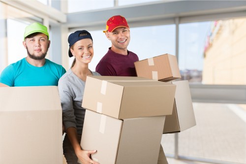 The image shows three professional house removalists inside a well-lit residential building, with large windows revealing a bright outdoor scene. The team, standing close together, is dressed in casual work attire with distinctively coloured caps: lime green, navy blue, and red. They are smiling and holding multiple cardboard boxes of various sizes, which are sealed with packing tape and appear to contain household items, possibly for a house move in Tooting or similar South London area. The boxes are made of brown kraft cardboard with visible flaps and taped edges. The background includes a corridor or living space with neutral-toned walls and a spacious atmosphere. The scene captures the organized and professional approach of Tooting Man with Van, a local removals service, as they prepare for a household relocation. The overall setting emphasizes the careful handling of belongings and teamwork involved in residential moving tasks.