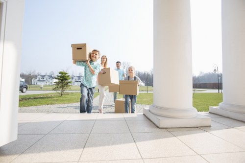 Four individuals, likely a family, are seen outside a residential building in Tooting, standing on a light-colored tiled porch framed by large white columns. One woman in a plaid shirt and jeans is in the foreground, smiling and holding a cardboard box above her head. Behind her, another woman in light clothing and glasses, along with a man and a young girl, are each carrying or holding cardboard boxes of various sizes, indicating they are in the process of moving. The boxes are plain brown and feature minimal markings, suggesting packing for a house removal or relocation. The background shows a suburban street with well-maintained lawns, small trees, parked cars, and a clear sky, signaling a typical day of a household move. The scene conveys a sense of organized activity associated with house removals, and the group’s casual attire and the presence of multiple boxes reflect the practical aspect of relocation services, such as those provided by Tooting Man with Van, in a local residential area near postcode SW17.