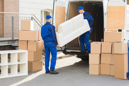 Two professional movers from Tooting Man with Van, dressed in blue uniforms with blue caps, are loading a white upholstered armchair into the rear of a large white van parked outside a residential building. The van door is open, revealing an interior designed for transporting household furniture. Surrounding the vehicle are several large cardboard boxes, some stacked to the left, others placed on the ground to the right, indicating a move or relocation in progress. A small white shelving unit with multiple compartments is also visible on the left side of the image. The scene is set on a paved driveway or street adjacent to a modern home with light-colored exterior walls, a metal railing, and steps leading up to the entrance. Natural daylight illuminates the scene, highlighting the textures of the cardboard, fabric upholstery, and the uniforms worn by the movers, all contributing to a professional and organized house removals operation in London, near postcode Tooting.