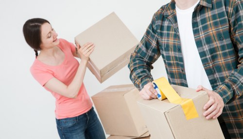 A woman in a pink top and jeans holding a cardboard box and smiling at a man dressed in a checkered shirt and white t-shirt, who is sealing a second cardboard box with yellow packing tape. They are standing in a bright, neutral indoor setting, possibly within a home or removal environment, with a plain light background. The scene depicts the process of packing or preparing belongings for a removal, fitting with services offered by Tooting Man with Van in the Tooting area near London, UK. The boxes are standard brown cardboard, and the woman appears engaged in arranging or checking the packed items, highlighting a typical house removals activity.