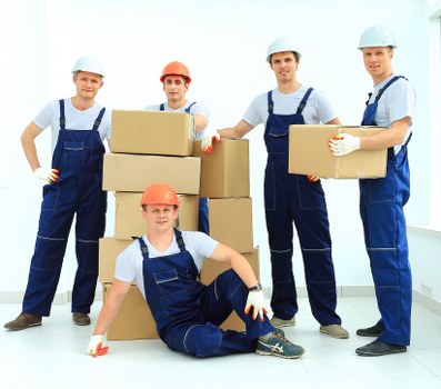 A group of five professional removal workers wearing blue overalls and safety helmets, with one worker in the front seated on the floor, poses inside a bright, modern residential space. They are surrounded by cardboard boxes, some stacked, which appear to contain household items. The Men with Van team is engaged in a house move, with the background showing a clean, white wall and a large window that allows natural light to illuminate the scene. The workers are holding or positioned near the boxes, ready to load or unload, and their expressions are friendly and professional. The setting suggests an organized and efficient household removal process in or around Tooting, South London, with the team prepared to assist with domestic relocations. The environment is neat, emphasizing the professional nature of Tooting Man with Van’s services, with a focus on house removals in the local area.