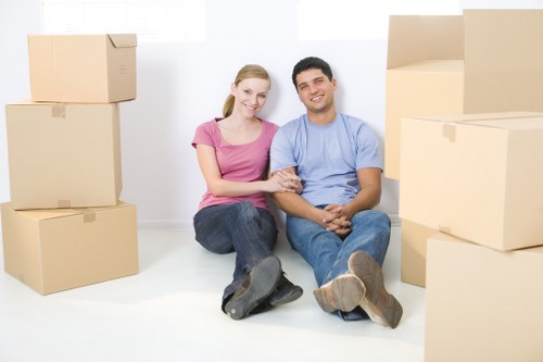 A young couple sitting on the floor in a room with white walls, surrounded by several large cardboard moving boxes of various sizes. The woman has blonde hair, wears a pink t-shirt and dark jeans, while the man has dark hair, wears a light blue t-shirt and jeans. They are smiling and appear relaxed, with the woman's arm linked through the man's, both sitting cross-legged. The scene suggests they are in the process of packing or preparing for a house move, which is consistent with professional removals services like those provided by Tooting Man with Van. The background includes no furniture or other objects, highlighting the focus on the packing process within a typical home environment, subtly referencing residential areas in Tooting, London.