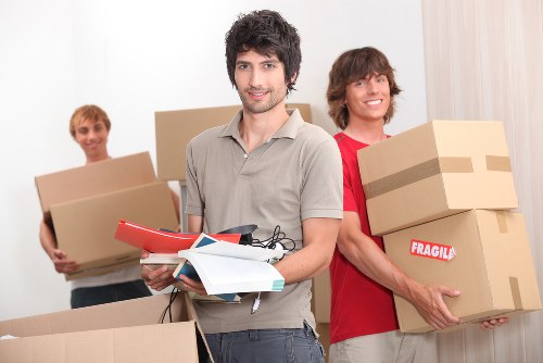 A group of three young men in a residential interior preparing for a house move, with one in the foreground holding other packing materials and a clipboard, and two others behind carrying cardboard boxes. The man in front has dark, slightly curly hair and is wearing a light grey shirt, while the two behind have lighter hair and are dressed in casual red and grey clothing. All are smiling. The boxes vary in size and are made of brown corrugated cardboard, some sealed with packing tape. One box held by the man in red is labeled with a red 'Fragile' sticker. The setting appears to be an internal space with a plain white wall and curtains visible in the background, indicating a typical house interior used for packing or moving activities. The overall scene reflects a professional removals service by Tooting Man with Van, aligning with house relocation tasks within London and nearby areas, such as Tooting or South West London postcode regions.