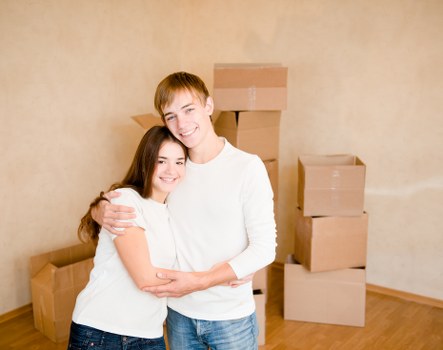 A young couple with light skin and brown hair, both wearing white tops, are smiling and embracing each other in a room with beige walls and wooden flooring. Behind them, there are several cardboard moving boxes of various sizes stacked against the wall, indicating a house relocation. Some boxes are fully closed, while others are open at the top. The scene appears to be inside a residential space, possibly a living room or hallway, with natural or soft lighting illuminating the area. The couple’s relaxed expressions and proximity suggest they are in the process of moving into or out of a home in Tooting or nearby south London. The setting conveys a moment of packing or unpacking during a house removal, which is typical for a professional removals service like Tooting Man with Van. Overall, the image captures a positive and organized atmosphere associated with residential moving within the local community.