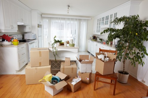 A spacious, light-filled kitchen with white cabinets and countertops, featuring a large window with sheer white curtains allowing natural light to illuminate the room. Several cardboard boxes are scattered across the wooden floor, some open revealing packing materials, kitchenware, and small household items. A small wooden chair is positioned to the right, next to a large potted plant with broad green leaves, adding a natural element to the scene. The kitchen includes various appliances such as a microwave, kettle, and toaster on the countertops, with some small potted plants and decorative items placed along the surface. The overall setting suggests an active house move, with packing boxes and household belongings prepared for transportation, typical of a property in the Tooting area, illustrating the services offered by Tooting Man with Van within the removals sector.