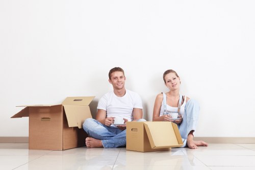 A young couple with light skin sitting on a light-coloured tiled floor inside a home, surrounded by open cardboard moving boxes. The man on the left is dressed in a white t-shirt and blue jeans, holding a tablet, and smiling at the camera. The woman on the right wears a white top and light blue jeans, holding a small box, with a neutral expression and slightly leaning against one of the boxes. Behind them, more cardboard boxes are stacked, some open and empty, and others filled with household items. The background features a plain white wall with no additional furnishings visible, indicating a simple interior space in a residential property, possibly in the Tooting or south-west London area. The scene suggests a house removal or packing process, typical of local removals services in the area, with natural lighting illuminating the scene and highlighting the textures of the cardboard, clothing, and clean tiled flooring.
