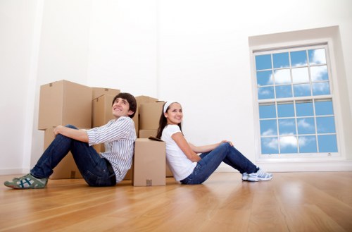 A young man and woman sit on the light wooden floor of an empty residential room with white walls, leaning against a stack of cardboard moving boxes of various sizes. The man, wearing a striped shirt and jeans, is smiling and looking upward, while the woman, in a white t-shirt and jeans, is also smiling and looking toward the camera. Behind them, there is a large double-glazed window with a white frame, showing a view of blue sky and clouds outside. The room appears to be part of a house in Tooting, with minimal furnishings and a bright, natural light environment, suggesting a recent move or packing process as part of a house removal service by Tooting Man with Van.