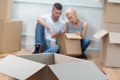 A young man and woman sit on the floor of a well-lit room surrounded by stacked cardboard boxes, some open and partially filled with household items. The man, wearing a white t-shirt and jeans, appears to be assisting the woman, who has short blonde hair and is dressed in a sleeveless top, as they prepare or pack items into a box. In the foreground, an empty, open cardboard box is prominently visible, while additional boxes are placed along the walls and on the floor in the background. The room has a neutral, contemporary interior with wooden flooring and a light-coloured wall, suggesting a residential setting typical of a house or flat in the Tooting area. The scene is calm and organized, reflecting activities involved in home removals, packing, or preparations for a move, with natural daylight illuminating the space, consistent with local house moving services like Tooting Man with Van.