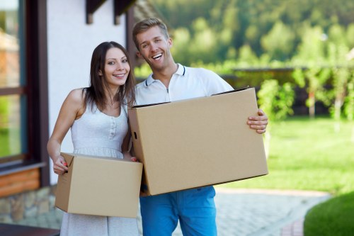 A smiling young couple stands outside a modern residential house during daylight, each holding a medium-sized cardboard moving box. The woman, with long dark hair, wears a sleeveless white dress and has a friendly expression, standing on the left. The man, with short light brown hair, is dressed in a light blue shirt and darker blue trousers, holding a larger cardboard box on his right side. They are positioned close together with their arms around each other’s waists. The background features a well-maintained garden with green grass, shrubs, and trees, as well as the exterior wall of a house with a white facade, brick accents, and large windows. The area appears bright with natural sunlight, suggesting a clear day, and the scene captures an outdoor moment typical of a house move, which is relevant to Tooting Man with Van's removals services in the Tooting area, south London. The overall scene emphasizes the completion or ongoing stage of a home relocation process with visible household items ready for transport.