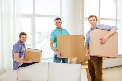 The image depicts three men in a bright, spacious living room with large windows allowing natural light to illuminate the space. The men are engaged in a house removal process, each holding or carrying cardboard boxes used for packing personal belongings. The man on the left, dressed in a purple shirt, is sitting on a white sofa and holding a medium-sized cardboard box with both hands. In the center, a man wearing a turquoise T-shirt is standing behind a white armchair and holding a larger cardboard box, smiling at the camera. The man on the right, dressed in a light blue shirt and brown trousers, is holding a sizable cardboard box, with one arm around the box and the other hand gripping its side, positioned near a white radiator and window frame. The room appears clean and modern, with neutral tones and minimal visible furniture and decor, consistent with professional house removals in the Tooting area, South London.