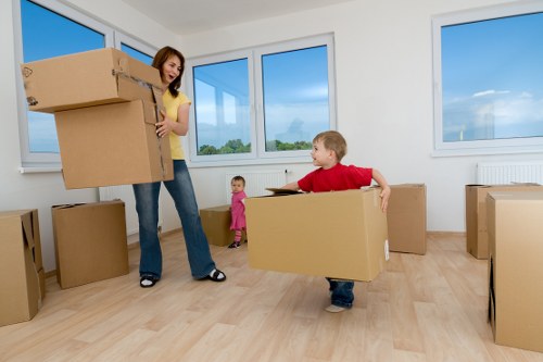 In a bright, spacious room with large windows showcasing a clear blue sky, a woman in casual clothing is seen holding a stack of cardboard boxes, likely preparing for a move in the Tooting area. The room has light wooden flooring and white walls, typical of a residential property in south-west London. A young boy dressed in a red shirt and dark trousers is actively carrying a medium-sized cardboard box, assisting with the relocation process, while a smaller child in a pink outfit stands nearby, observing. Several other cardboard boxes of various sizes are placed around the room, some stacked on the floor and others ready to be moved. The scene captures a moment of family collaboration during house removals, with the focus on packing or unpacking items in a typical home environment, emphasizing the practicality and familiarity of residential moving services offered by Tooting Man with Van. The overall atmosphere is well-lit with natural daylight, highlighting the clean and organized interior setup suitable for professional house removals and local relocation tasks in the Tooting postcode area.