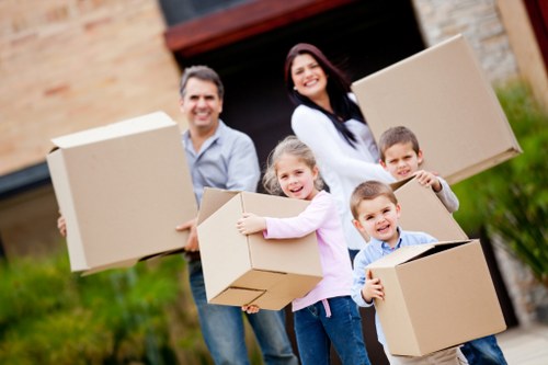 A family of four, including a father, mother, and two young children, standing outside a residential house on a suburban street near Tooting, South London, holding medium-sized cardboard boxes during a house move. The father, wearing a light blue shirt and jeans, is positioned to the left and smiles at the camera while holding a box with his right hand. The mother, with long dark hair and a white top, is on the right, also smiling, holding a box in both hands. The two children, a girl and a boy, are in front of their parents; the girl, wearing a pink sweater, hugs a box close to her chest, and the boy, in a striped shirt, holds another box, both facing the camera with smiles. The background shows part of the house’s brick facade, a driveway, and a garage. The green lawn and garden plants are visible behind the family, and the scene is illuminated by natural daylight, indicating a clear, pleasant day. This image is relevant to house removals services offered by Tooting Man with Van in the local area.