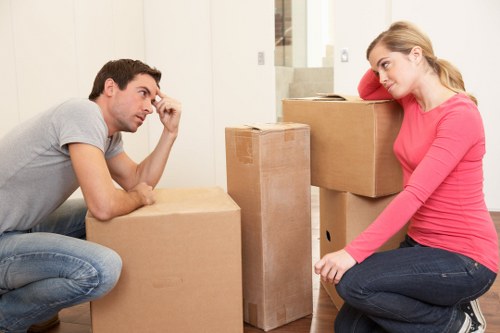 In a well-lit, neutral-toned interior space, a man and woman engaged in a discussion related to house removals are present. The man, with short dark hair and dressed in a light grey T-shirt and jeans, is seated on the floor, leaning onto a cardboard box, with a thoughtful expression and his hand resting on his forehead. The woman, with long blonde hair tied back and wearing a pink top and dark jeans, sits cross-legged on the floor, leaning against a large cardboard box, appearing to listen attentively. Surrounding them are several variously sized cardboard moving boxes, some sealed with tape and others partly open, containing household items. The boxes are positioned on a light-colored wooden floor and stacked on smaller units near a modern staircase with a simple white railing, suggesting a typical residential setting in Tooting or the surrounding London area. The scene depicts preparations for a move, with the focus on furniture and belongings such as boxes, ready for transportation by a house removals service like Tooting Man with Van. The natural lighting and minimal background detail support the context of an interior room during a house relocation process.