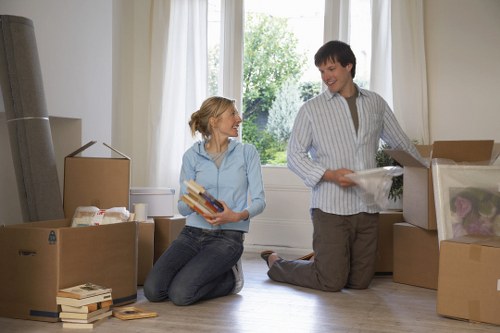 A woman with blonde hair tied back, wearing a light blue shirt and jeans, is kneeling on a wooden floor inside a house, holding a stack of books and smiling at a younger man with short dark hair. The man, dressed in a light grey striped shirt and dark trousers, is kneeling beside open cardboard boxes filled with household items such as clothing and decorative objects. The scene takes place in a bright room with large front-facing windows, allowing natural light to illuminate the space. Outside the window is a view of a residential street with greenery. The room appears to be a living area, with additional packed boxes stacked nearby, some sealed and others open. The overall environment reflects a domestic setting during a house removal or relocation process, with a focus on packing and organizing belongings, as part of a professional removals service in Tooting or the surrounding area, associated with Tooting Man with Van, according to the context of the website page on insurance and safety.