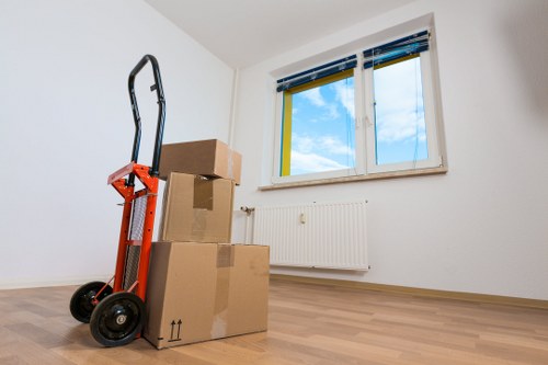 An interior room with white walls and a light wooden floor, featuring two large cardboard boxes stacked on top of each other, with a smaller box in front. The boxes are sealed with brown packing tape and appear to contain household items suitable for a house move. To the left of the boxes is a red hand truck with black wheels, positioned upright and leaning slightly against the boxes, ready for lifting or transportation. The room has a large double-glazed window with a white frame, revealing a clear blue sky with some clouds and a partial view of neighboring buildings outside. Below the window is a white radiator attached to the wall. The scene suggests preparations for moving or packing within a residential property in Tooting, London, with natural daylight illuminating the space and highlighting the clean, minimal decor typical of a modern house or flat.