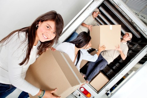 A smiling woman with long brown hair, dressed in a white top, is holding a medium-sized cardboard box in a home removal setting, standing in front of an open removals van. Inside and around the van, two men are visible; one is holding a large cardboard box near the van's entrance, wearing a light-colored shirt and dark trousers, while the other, dressed in a grey top and dark trousers, is carrying a box into the vehicle. The interior of the van shows several more boxes arranged in the cargo area, suggesting an organized move. The scene is set on a residential street with the rear hatch of the van open, revealing the loading process. The boxes vary in size, with some appearing to be packed with household items while others are empty or lightly packed. The environment is well-lit, likely by natural daylight, providing clear visibility of all objects, textures, and colors, which include cardboard boxes, soft fabric clothing, and the metal and plastic structure of the removal vehicle. The scene exemplifies professional house removals typical of Tooting, South West London, and features the local removals company, Tooting Man with Van, actively engaged in a stress-free, organized move. Overall, the image captures the orderly process, the teamwork involved, and the typical equipment used in a residential relocation in the area. Detailed descriptions of household items and the professional approach support the site's SEO and accessibility goals for a house removals service website.