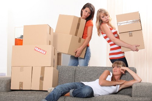 Two young women are in a living room filled with moving boxes, preparing for a house relocation. The woman on the left, dressed in a red top and jeans, is carrying a medium-sized cardboard box, which appears to be packed and sealed with packing tape. The woman on the right, wearing a red and white striped dress, is holding another cardboard box, slightly smaller, with visible packing tape securing its flaps. In the foreground, a young boy with blond hair, wearing a white T-shirt and blue jeans, is lying comfortably on a grey fabric sofa, resting his head on his hand and looking off to the side. The background features a white wall, and the scene suggests an interior residential space typical of a house move in the Tooting area or broader south London location. The boxes vary in size and are stacked on the sofa and the floor, some labeled with fragile stickers, indicating careful household packing. The natural lighting and casual atmosphere reflect a typical domestic moving scenario, involving household furniture and personal belongings ready for transport, with a focus on local house removal services like those provided by Tooting Man with Van.