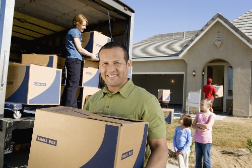 In the foreground, a smiling man in a green shirt, holding a medium-sized cardboard box labeled 'Small Box,' stands outside a suburban house on a residential street, possibly in Tooting or nearby. Behind him, a young woman unloads several similar labeled cardboard boxes from the back of a large moving van parked on the driveway. The van's open side reveals a partially filled interior with more boxes inside. To the right, three children stand on the grass near the sidewalk, observing the activity; one girl in a pink top and blue jeans, another girl in a pink top and brown pants, and a third child in a blue top and shorts. The house features a grey tiled roof, a front porch with an arched opening, and a closed garage door. The scene is lit by daylight, indicating a clear, sunny day. This setting depicts a typical house removal process managed by Tooting Man with Van, highlighting the careful handling of household items during a relocation within the local area, emphasizing professionalism and household furniture management during a house move in south-west London.