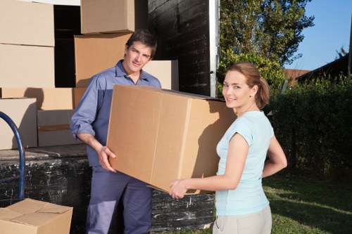 A young man and woman are standing outdoors in front of a black removals van, with the man handing over a large cardboard box to the woman. The man is dressed in a blue shirt and dark trousers, while the woman is wearing a light blue T-shirt and beige trousers. The van's rear doors are open, revealing multiple stacked cardboard boxes and packages behind them, some partially visible. The scene takes place on a residential street with green trees and bushes visible in the background, under clear daylight. The cardboard boxes vary in size, some with visible flaps, and are used for packing personal belongings during a house move. The individuals appear to be engaged in a coordinated move, likely organized by Tooting Man with Van, a professional house removals company operating in the Tooting area. The overall setting emphasizes the logistics of packing and moving household items, with a focus on sustainability and recycling, aligning with the webpage about recycling and sustainability in house removals.