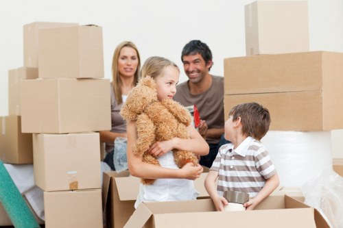 A family of four preparing for a house move inside a bright, modern room, surrounded by numerous cardboard boxes of various sizes, some sealed with packing tape and others open, revealing household items such as small appliances and personal belongings. The mother, with long blonde hair, stands behind the children, smiling gently. The father, with short dark hair, appears to be engaging with the children, also smiling. In the foreground, a young girl with blonde hair dressed in a beige, fluffy sweater holds a small teddy bear, while a boy in a striped shirt sits inside an open cardboard box, holding a metal container. The scene is well-lit with natural daylight, indicating daylight hours, and the background shows a plain white wall, suggesting an indoor residential setting in Tooting, London. The overall atmosphere captures the preparatory stage of a house removal undertaken by Tooting Man with Van, part of their removals services in the South London area, with the focus on packing, family involvement, and household relocation activities.